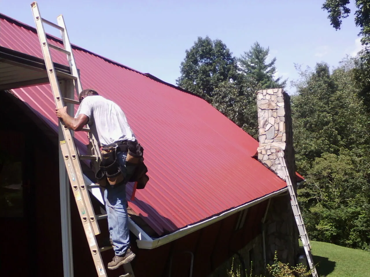 Roofer on a ladder beside a red metal roof and stone chimney — Soffit & Fascia Repair by Roofing Brothers in Bow